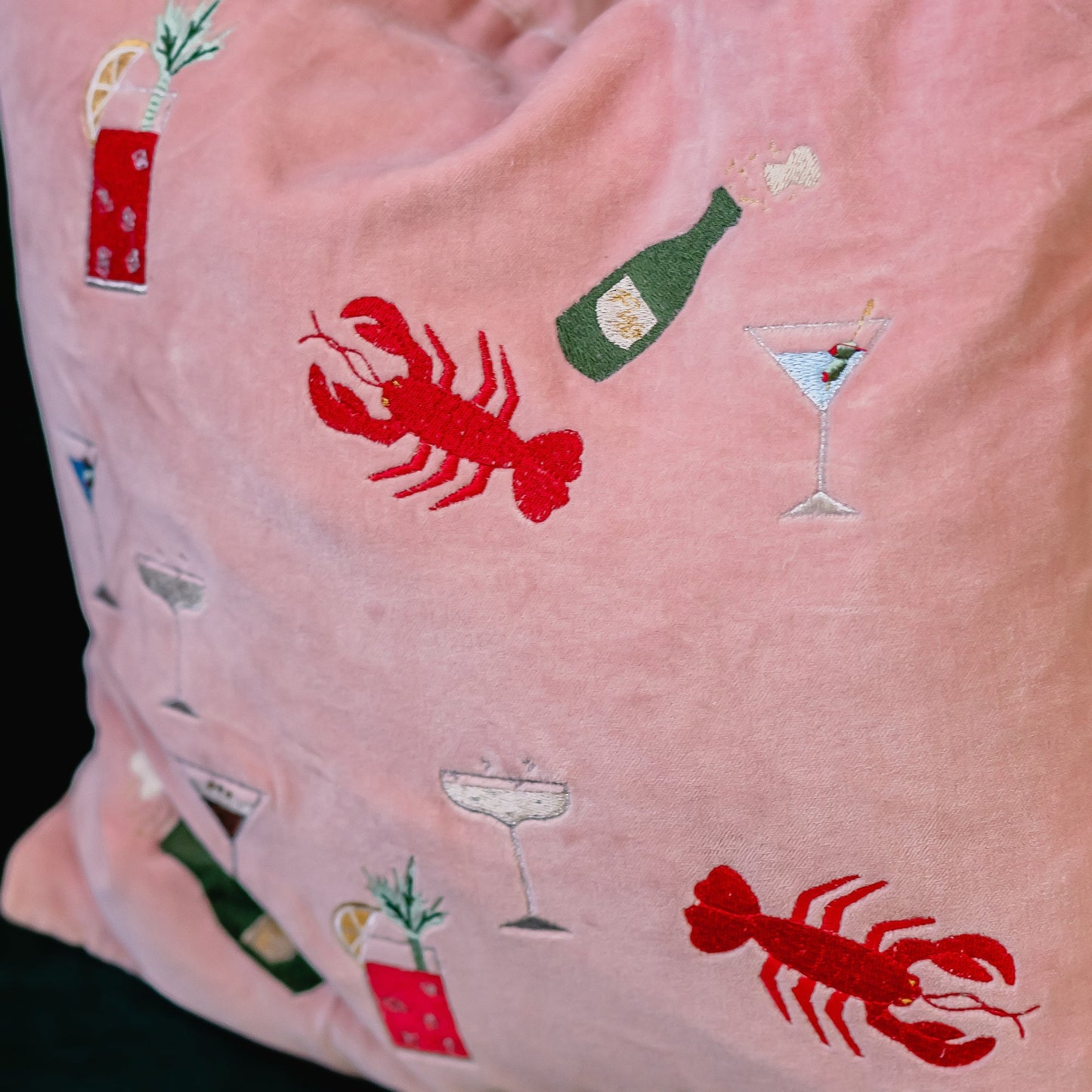 A pink velvet cushion with embroidered lobsters, champagne and martinis on it