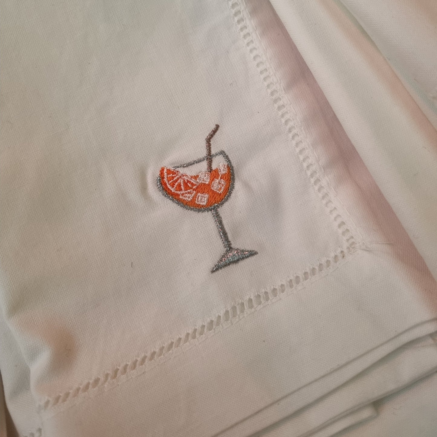 A white cotton napkin with an embroidered design of an orange-colored spritz cocktail glass, complete with a metallic straw.