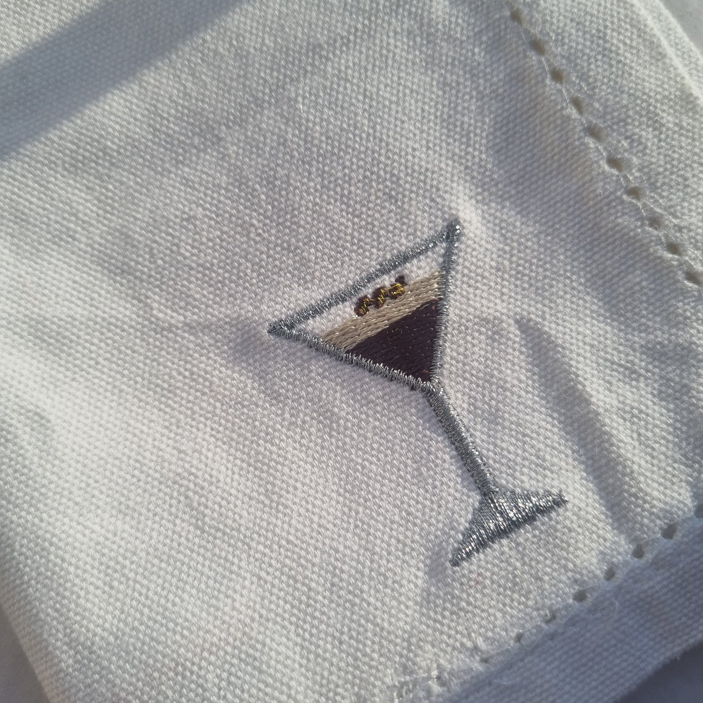 A white cloth napkin with a silver embroidered design of an espresso martini cocktail