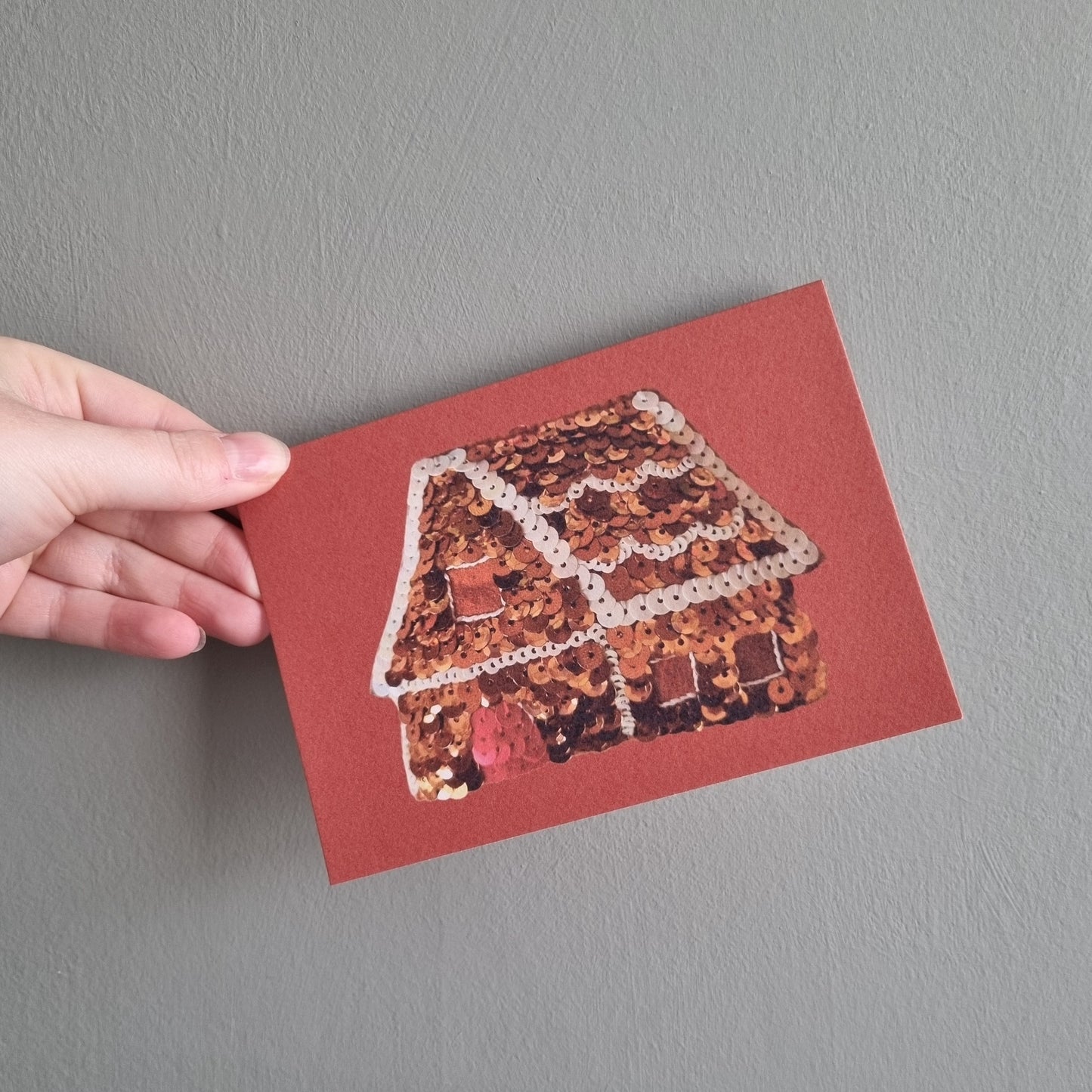 Kate's hand holding a red card with an image of a gingerbread house made of sequins on it
