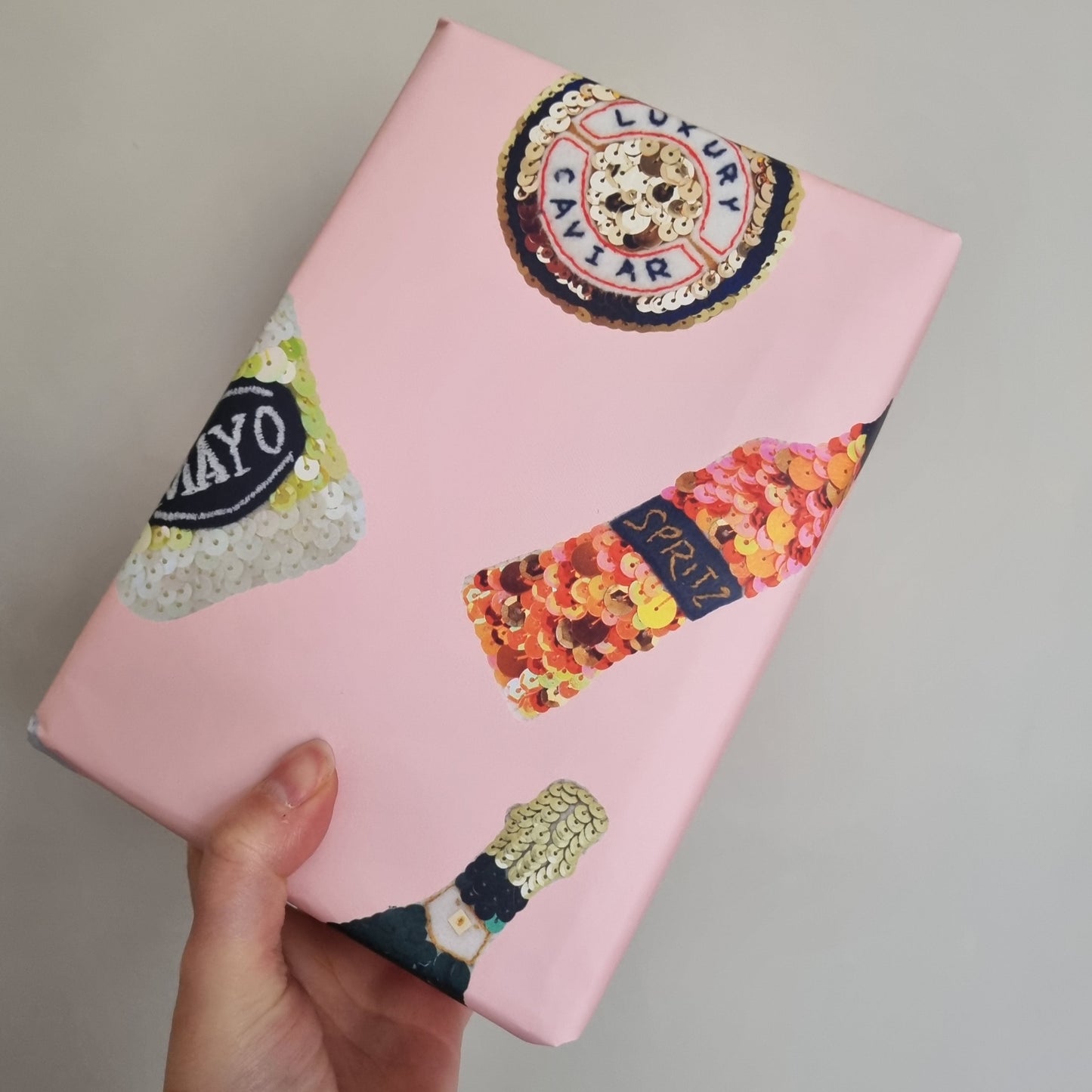 Kate's hand holding a small package covered in her pink wrapping paper with printed images of sparkly sequin ornament on it