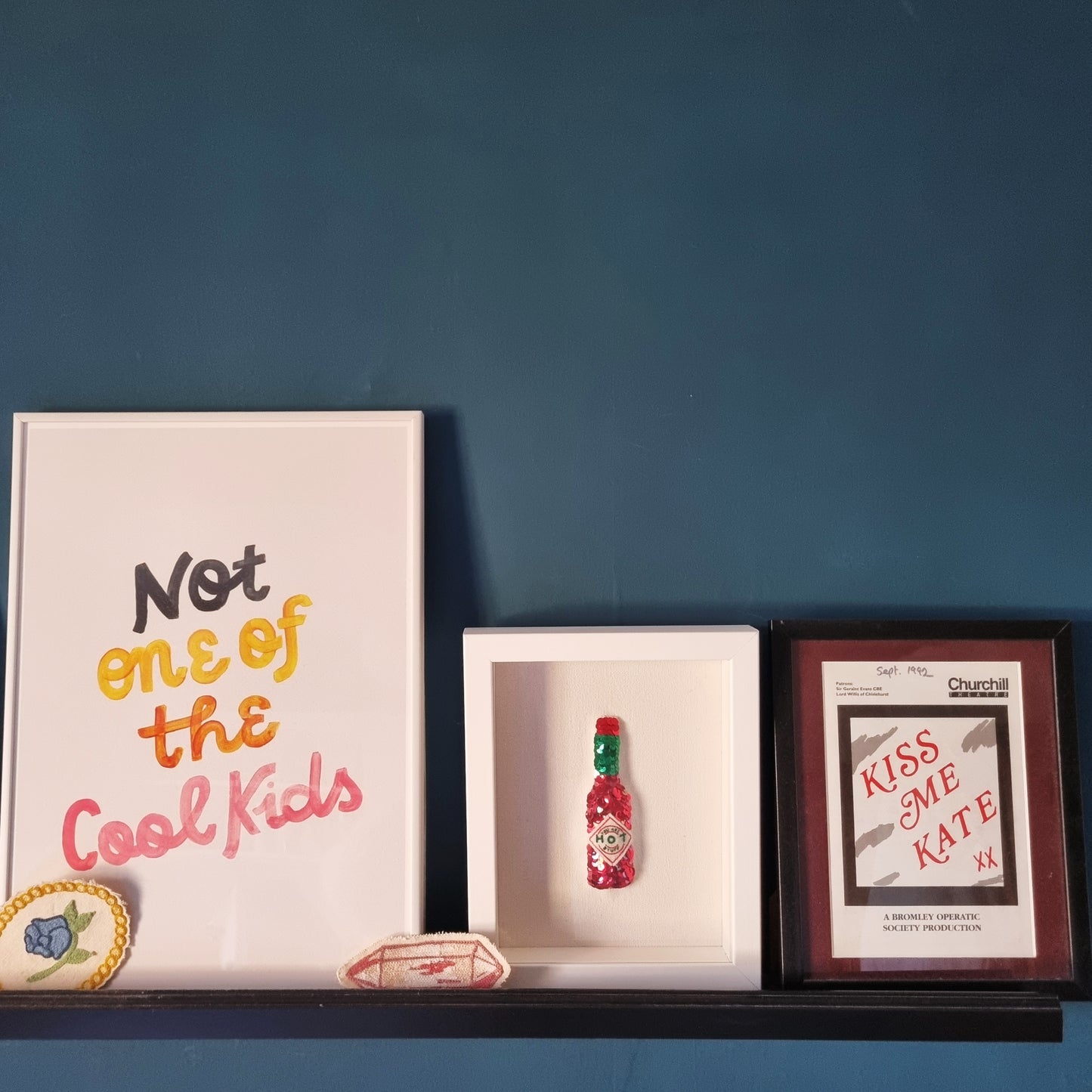 The original sequinned tabasco sauce bottle saying you are hot stuff, framed in a white box frame on a black shelf surrounded by other art