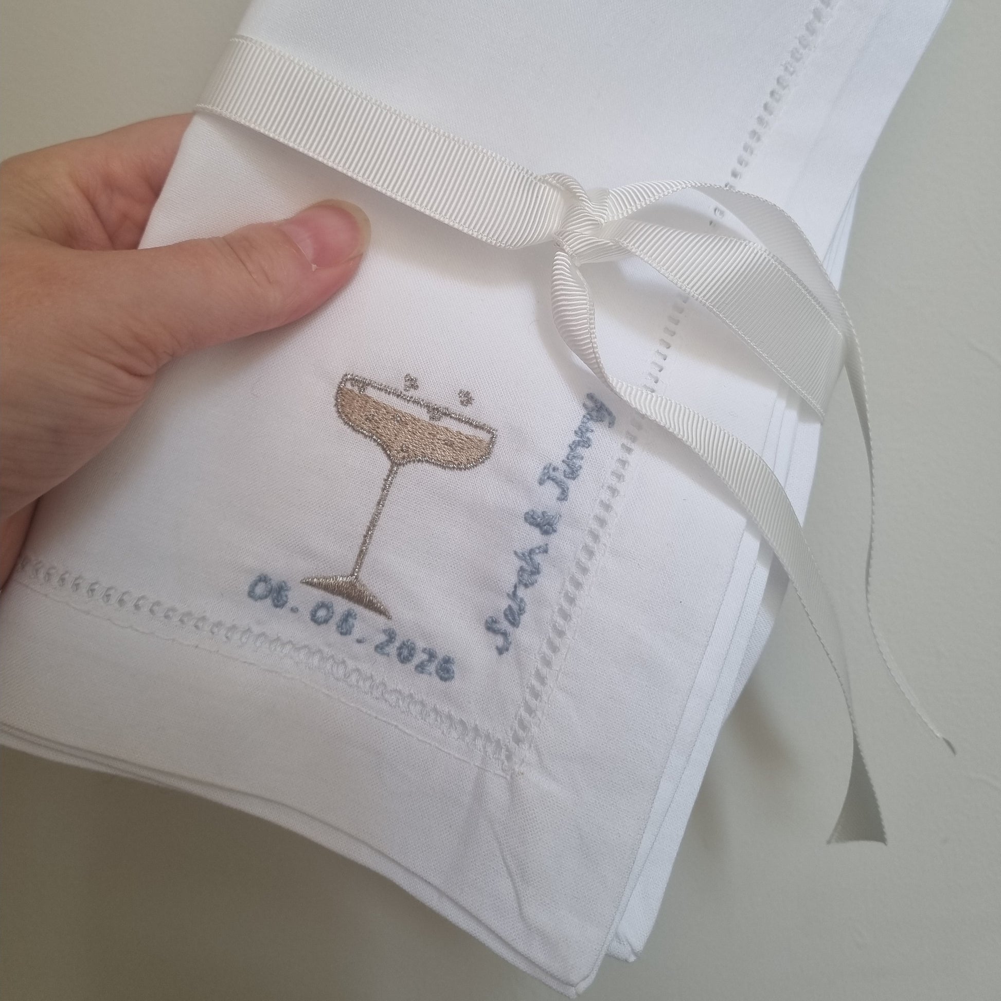 A close up of two napkins with embroidered champagne glasses and personalised with blue embroidery with names and dates