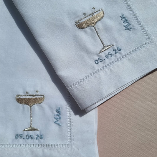 Two folded white embroidered table linens with stitched borders, gold champagne coupe glass motifs, and hand embroidered blue personalised names and dates,