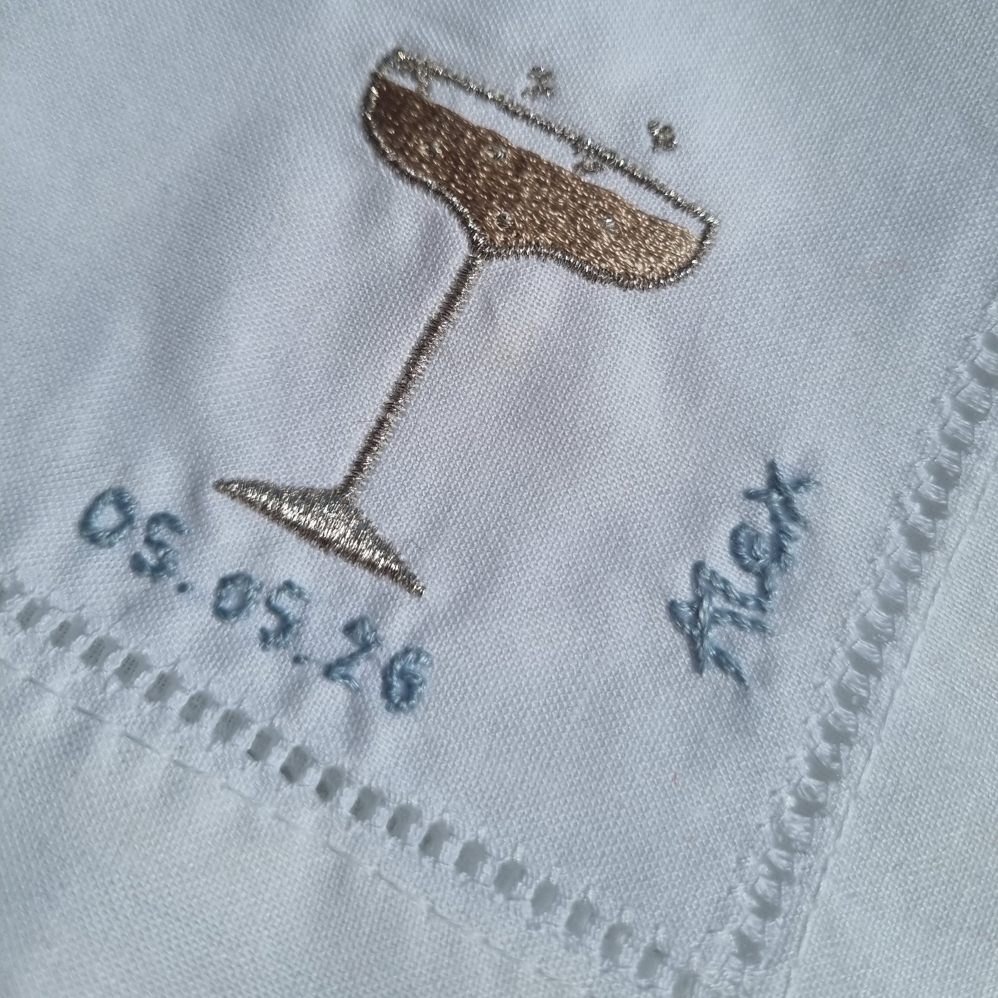 Close-up of a white embroidered fabric with a metallic champagne coupe design, blue stitched border, and blue embroidered text.