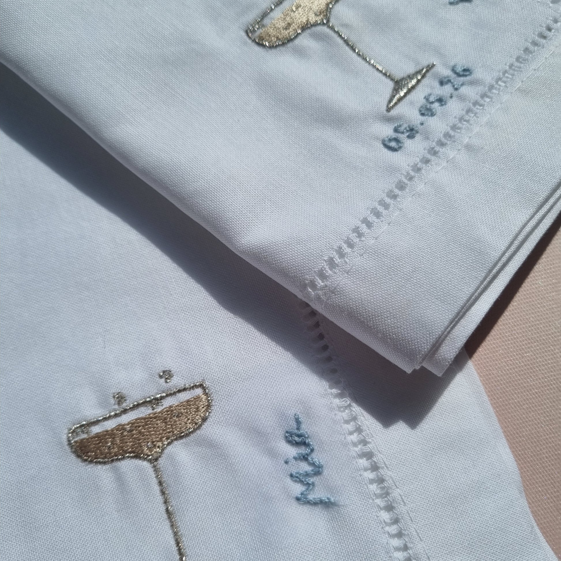 Close-up of folded white napkins with embroidered golden champagne glass motif plus blue hand  stitched name and monogram details.