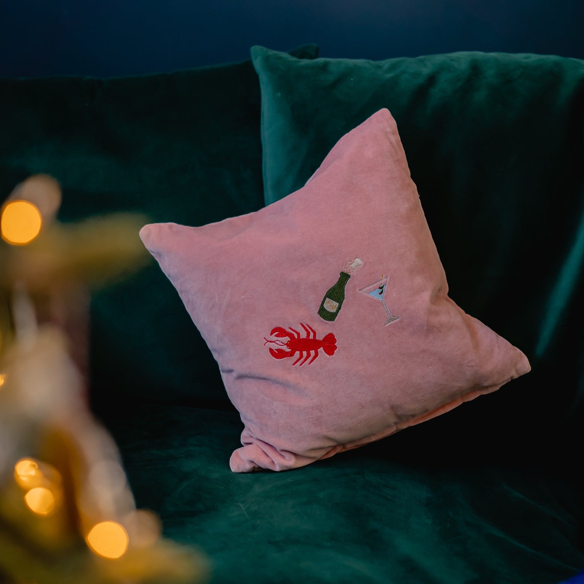 A pink velvet cushion with an embroidered Lobster, Champagne Bottle and Martini  by Kate Gwilliam, Queen Of Sequins