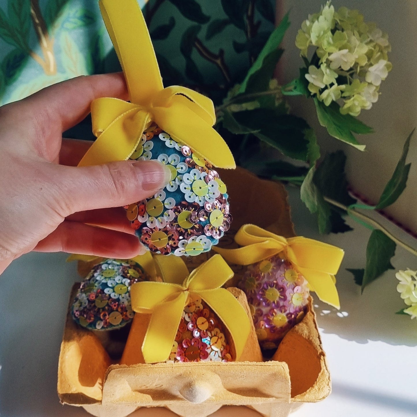Kate's hand holding a blue sequin egg above a box of sequin eggs