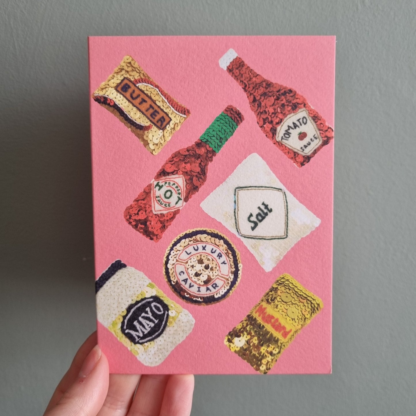 Kate's hand holding a pink card covered with images of condiments from original embroideries