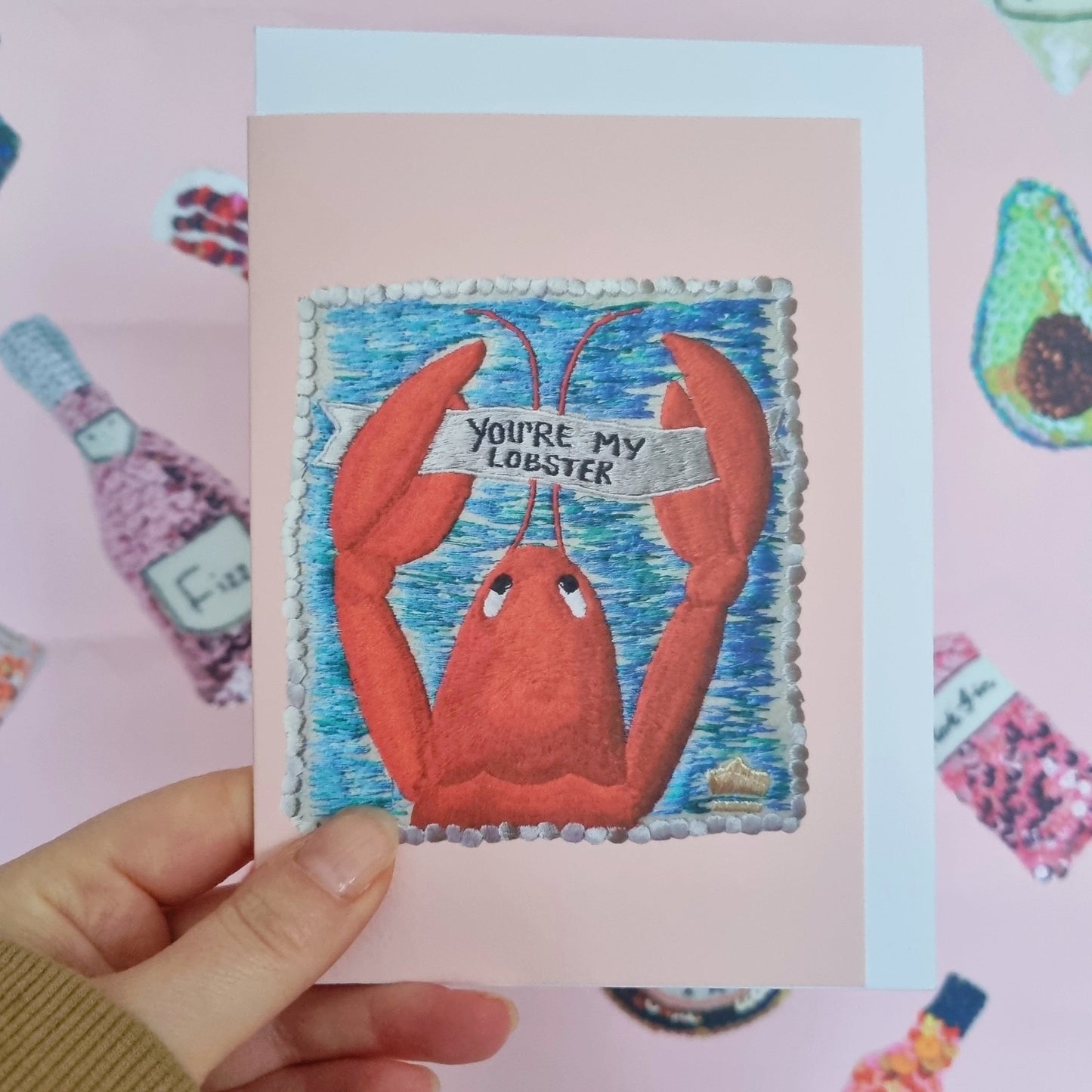 Kate's hand holding a pale pink card with an image of an embroidered lobster holding a banner saying "You're my Lobster"