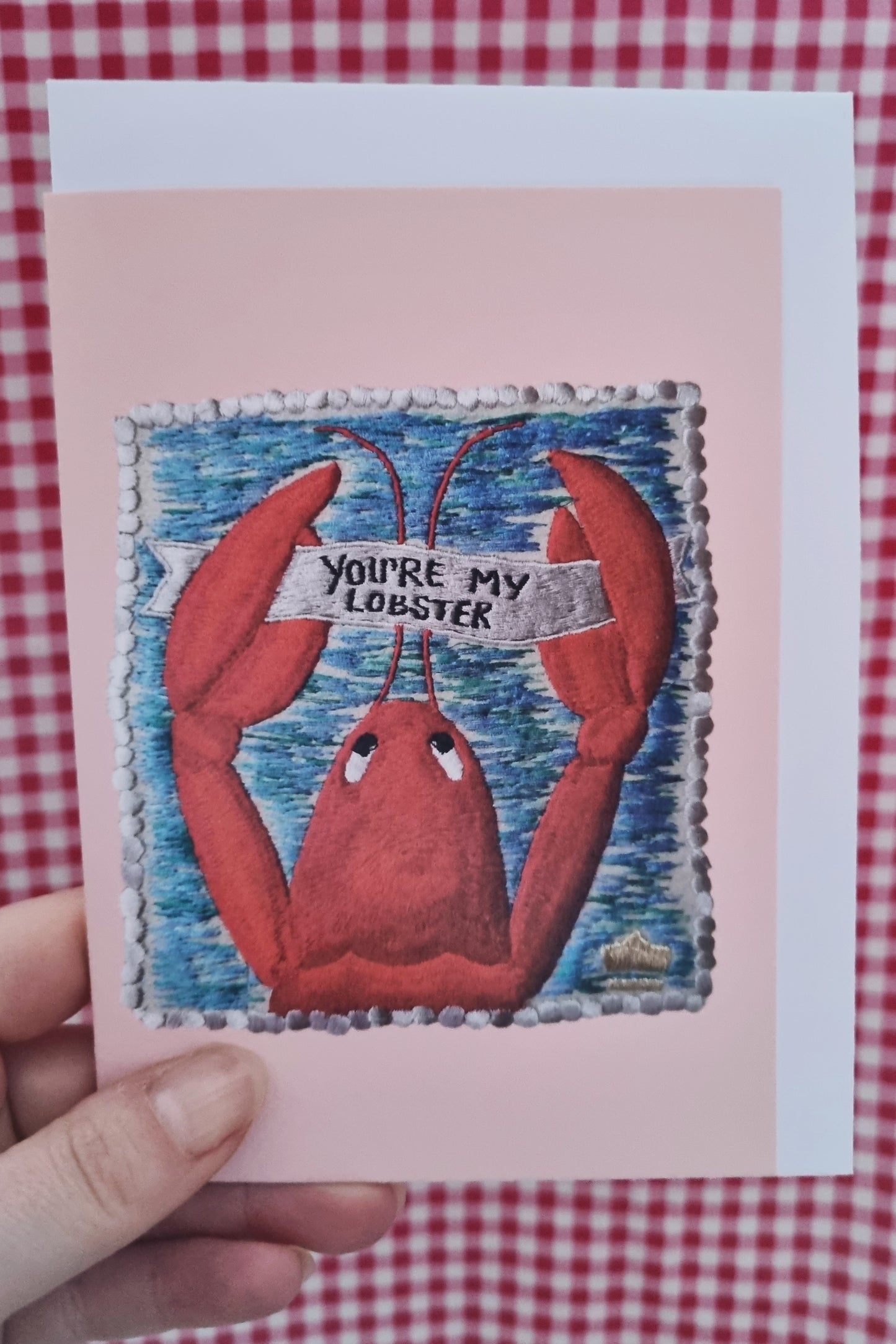 Kate's hand holding a pink card with an image of an embroidered lobster stampf