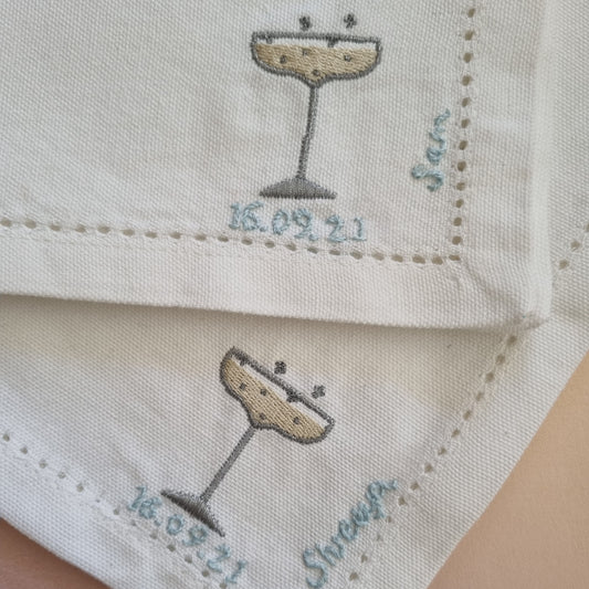 A close up of two napkins with embroidered champagne glasses and personalised with blue embroidery with names and dates