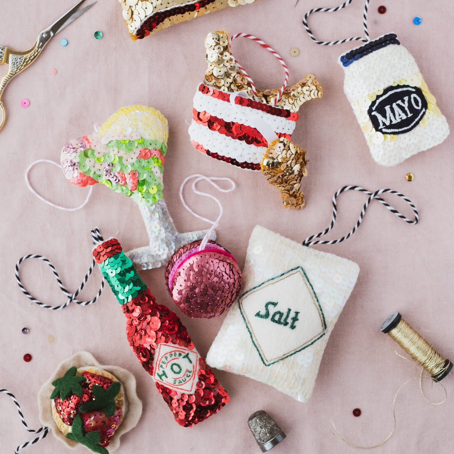 A collection of sequin ornaments on a pink background including a pink macaron, tabasco sauce bottle, fried chicken bucket and mayo jar